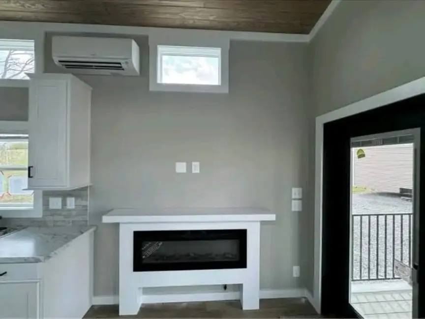 Modern room with a wall-mounted air conditioner, small window, and electric fireplace. Large door on the right opens to a porch. Bright and minimalist.