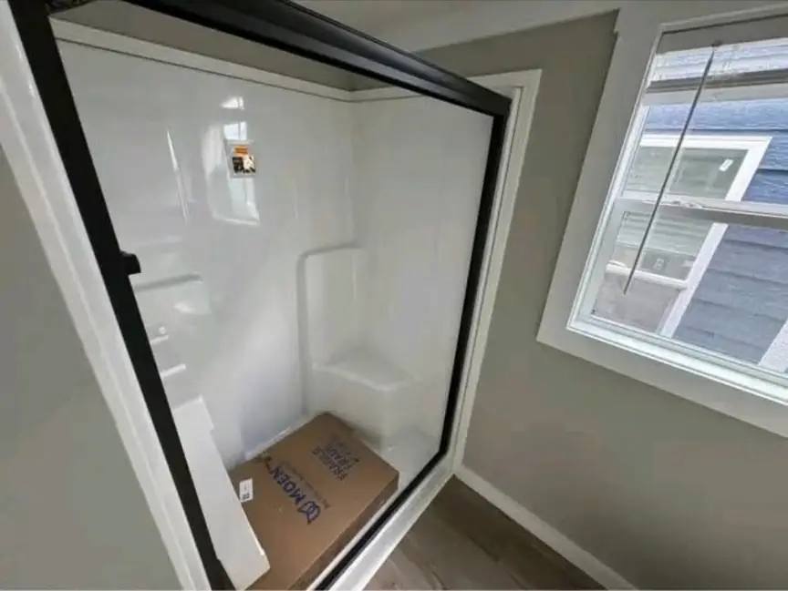 A white shower stall is installed backward, facing a room instead of a bathroom. A box is inside, and a window is on the right wall, creating a humorous tone.