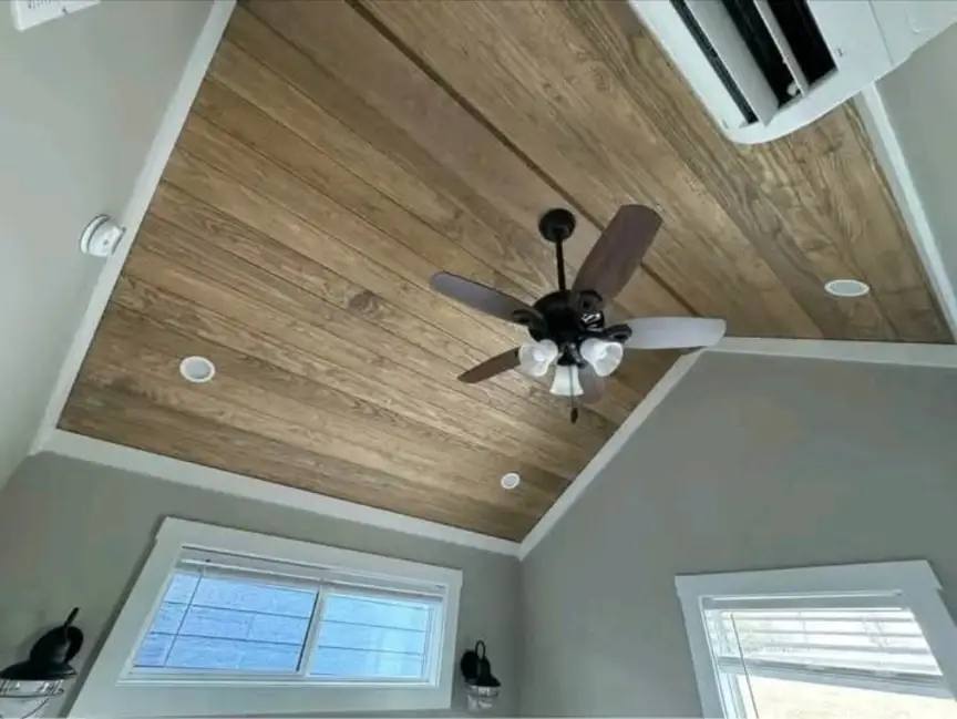 Wooden ceiling with a central ceiling fan featuring five blades and three lights. Two windows with blinds and a mounted air conditioner are visible.