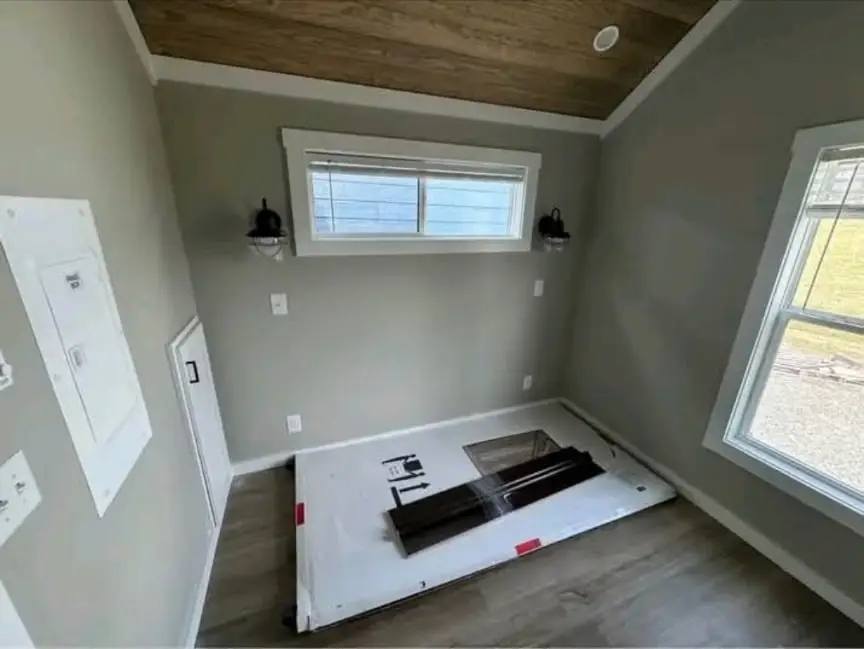 Small gray room with a sloped wooden ceiling and two windows, one horizontal and one vertical. A cardboard box lies on the floor, suggesting new furniture.