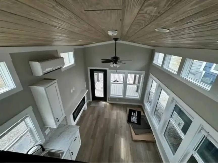 Aerial view of a modern, compact living room with light wood floors and a wooden ceiling. Large windows and a black ceiling fan add an airy feel.