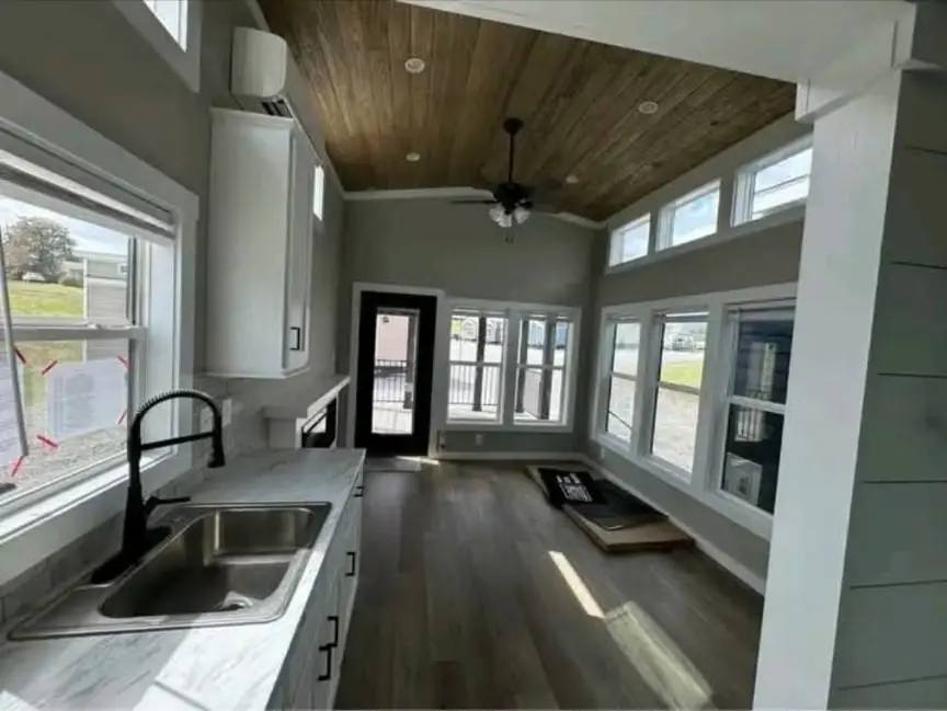 A modern, narrow room with large windows, hardwood floors, and a marble countertop with a black faucet. The ceiling is wood-paneled with a ceiling fan.