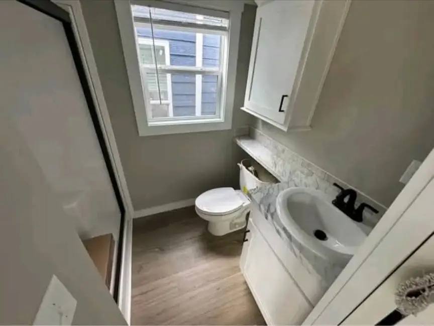Narrow bathroom with light wood floor, white walls, a small window, a white toilet, and a white sink with marble countertop and black faucet.