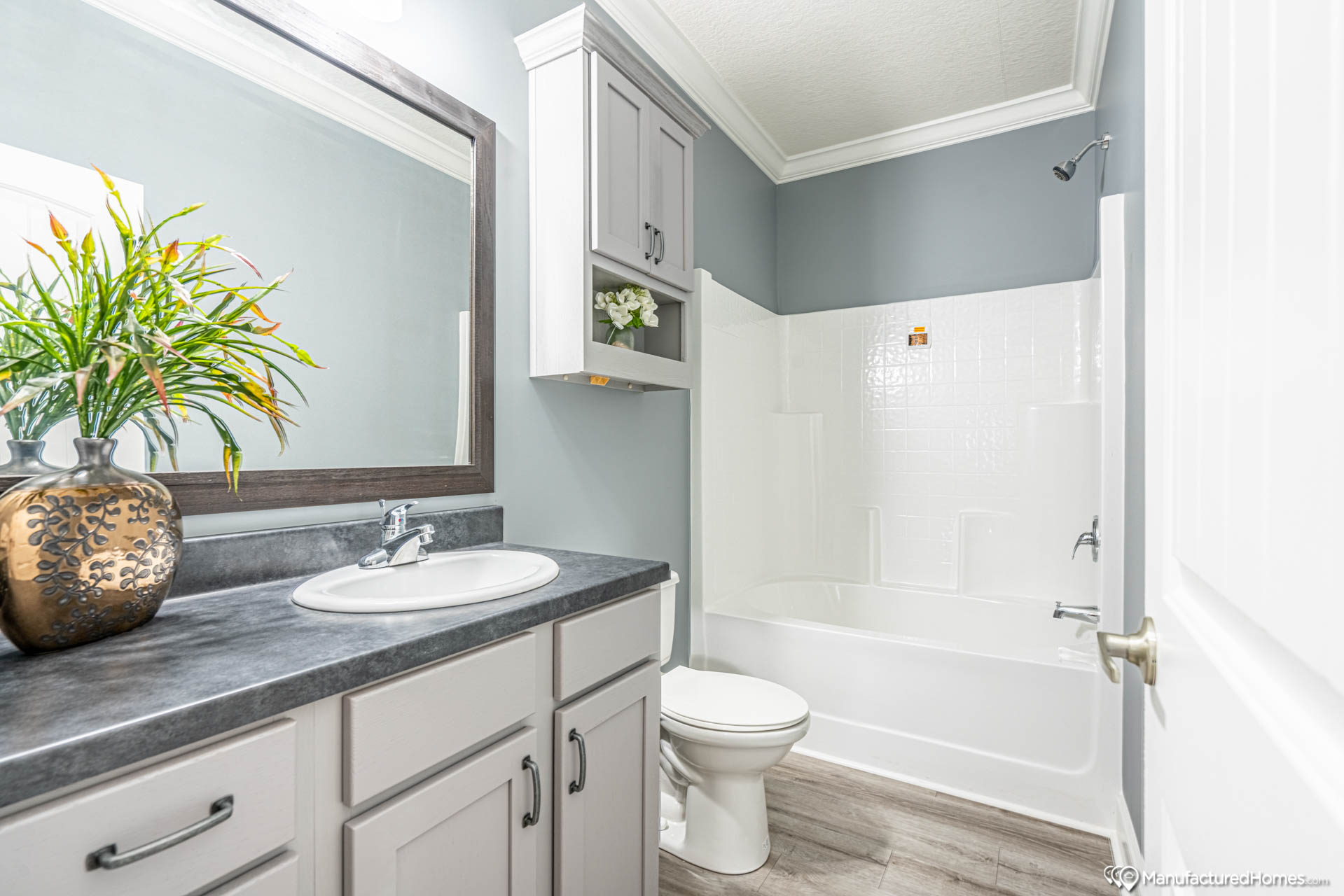A modern bathroom with a gray countertop, white sink, and cabinets. A large mirror reflects light, and a potted plant adds vibrancy. The white tub and tiled shower are neatly arranged, conveying a clean, serene atmosphere.