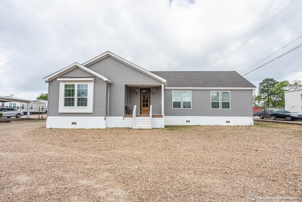 A single-story gray house with white trim sits on a gravel lot. It features a small front porch with steps and large windows, set against a cloudy sky.