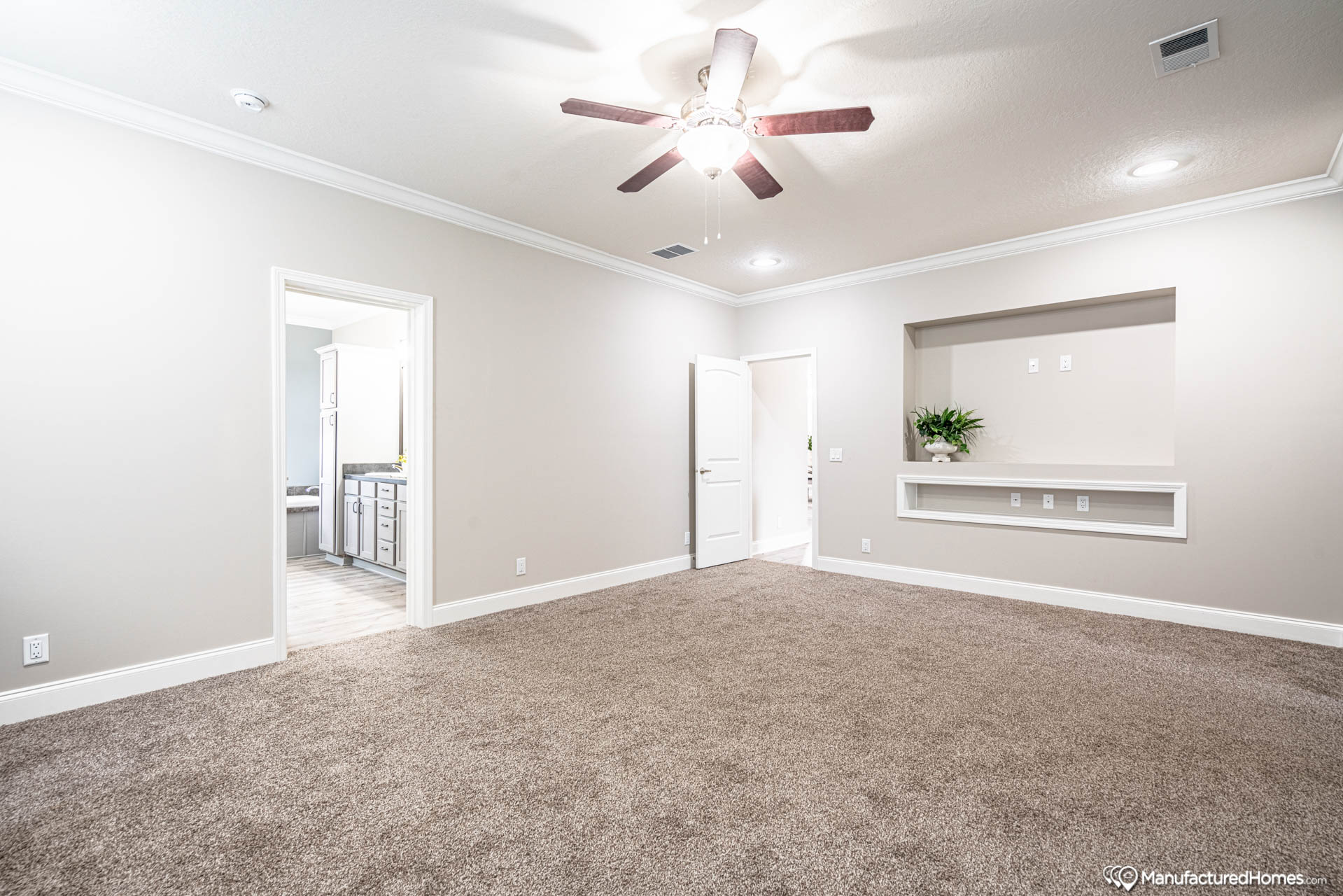 A spacious, empty room with beige walls and carpeted floors. A ceiling fan with lights centers the ceiling. A doorway leads to a bright room with cabinets, and a built-in shelf holds a green plant for a minimalist feel.