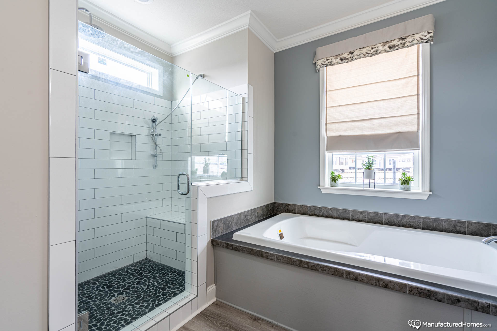 A modern bathroom features a glass-enclosed shower with white subway tiles and pebble flooring, adjacent to a white bathtub under a window with a beige shade.