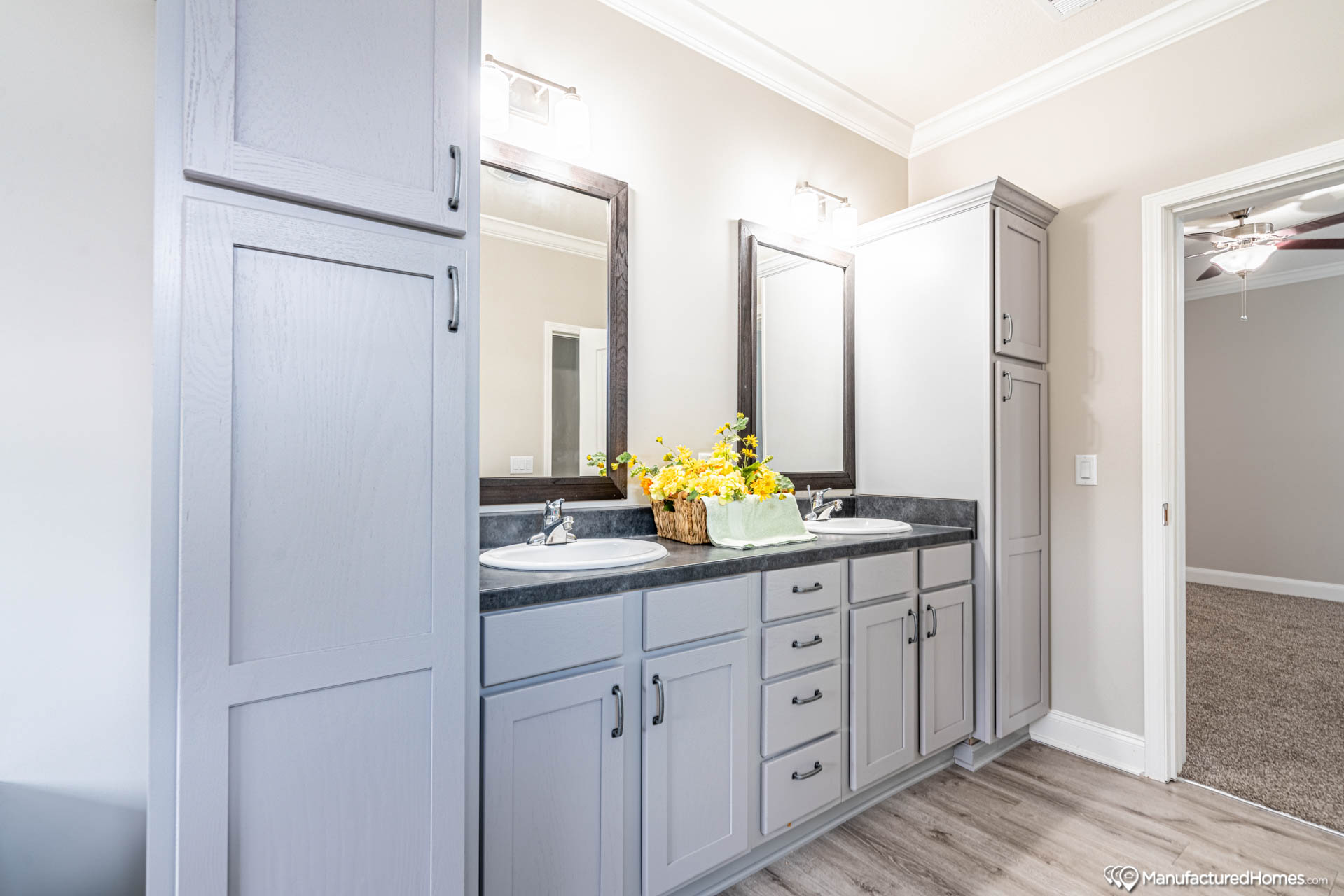 A bright bathroom with light gray cabinets, dual sinks, and black countertops. Two mirrors above the sinks, adorned with yellow flowers in a basket.