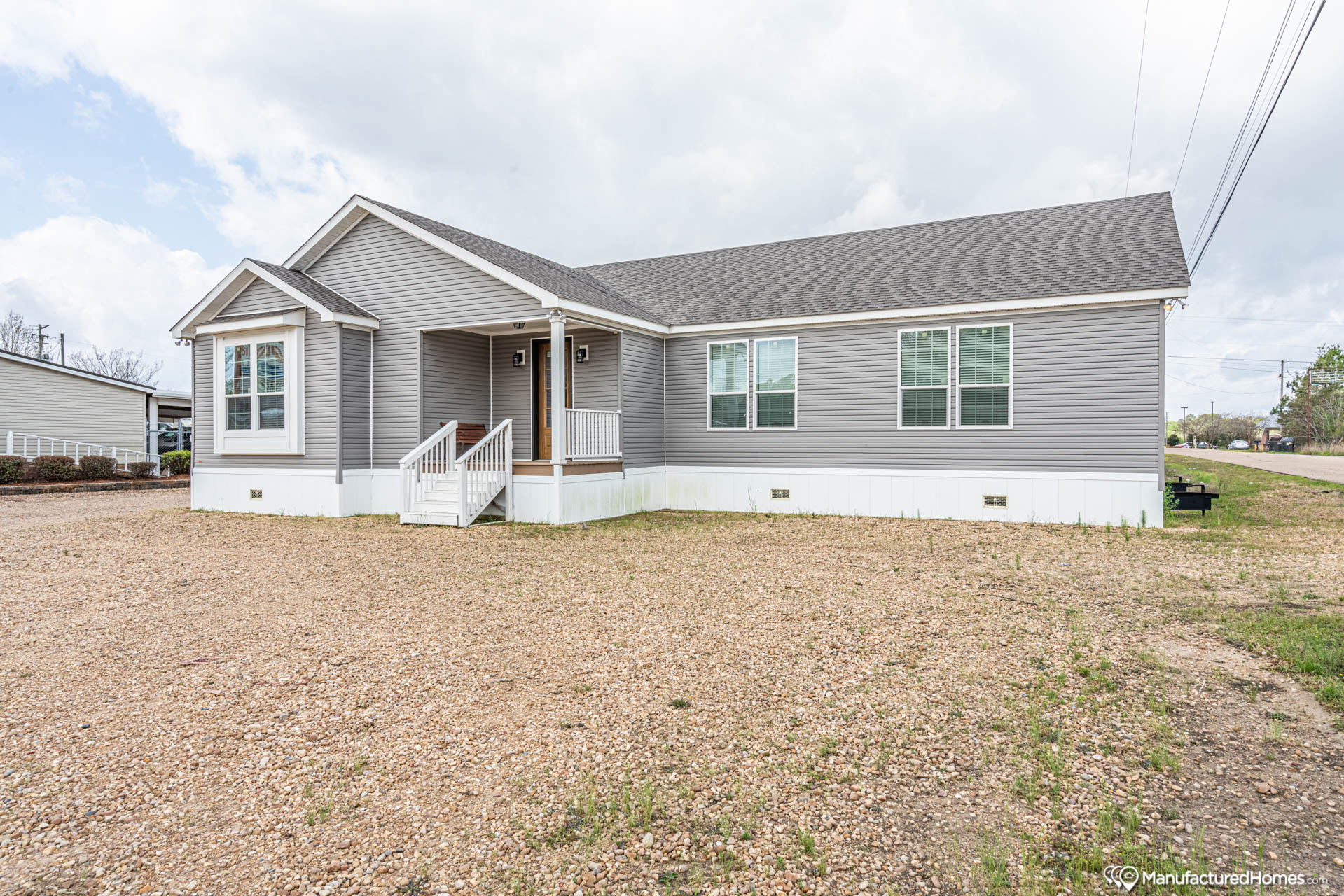 A gray manufactured home with white trim, featuring a small porch and steps. It sits on a gravel driveway under a cloudy sky, conveying a suburban setting.