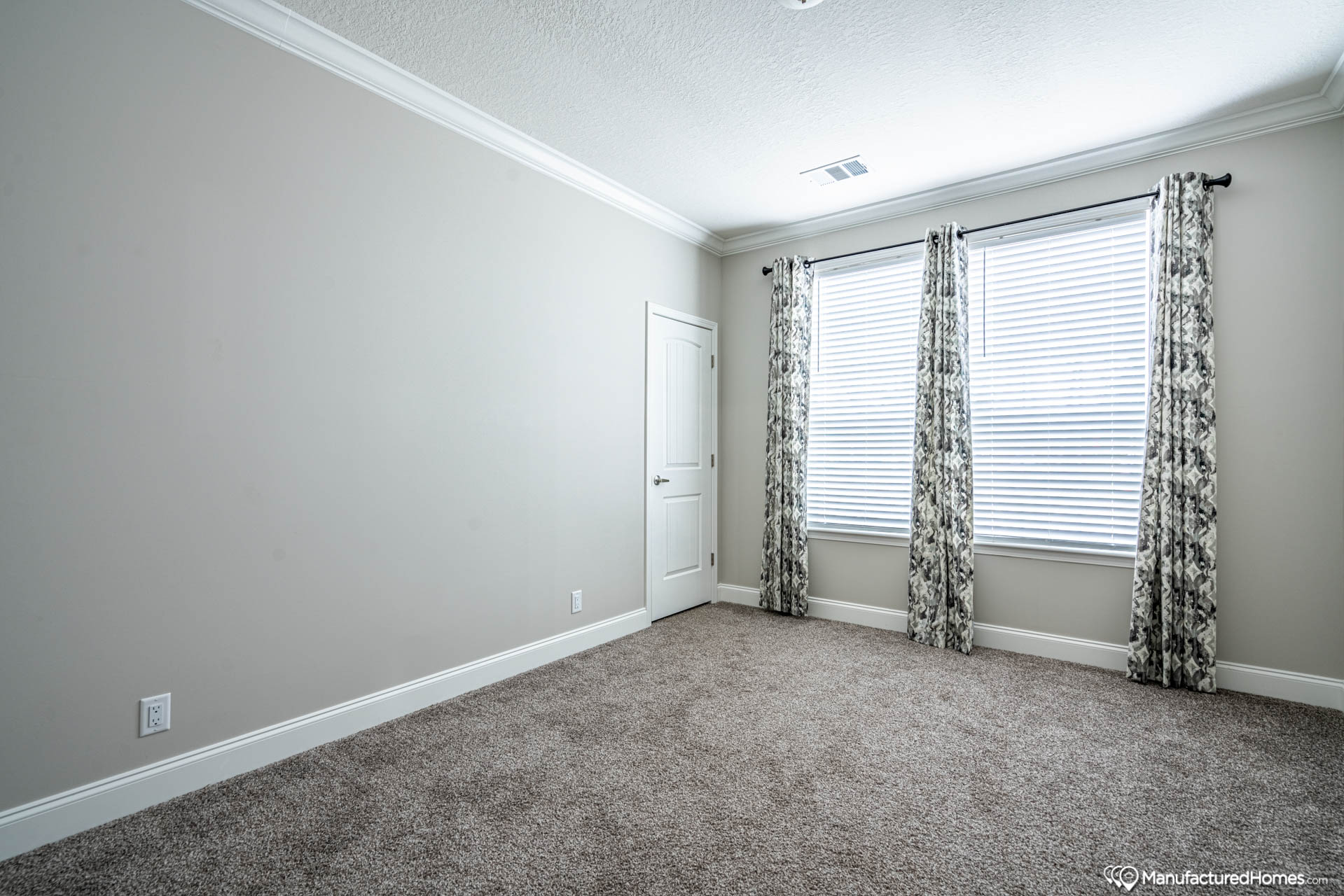 An empty room features beige walls, a white door, and two large windows with floral curtains. The carpeted floor and soft lighting create a tranquil, minimalist atmosphere.