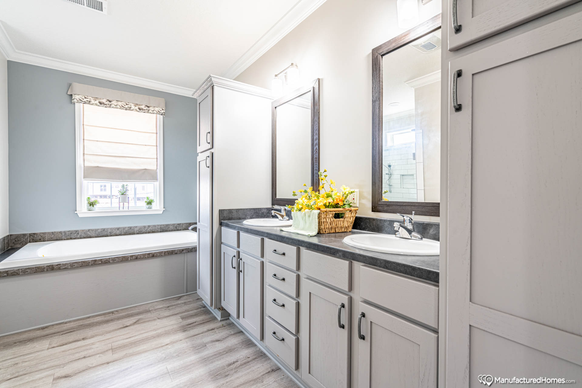 A modern bathroom with a dual-sink vanity, gray cabinets, and a large mirror. A basket of yellow flowers adds a cheerful touch. A soaking tub sits near a window, and the room has light wood flooring.