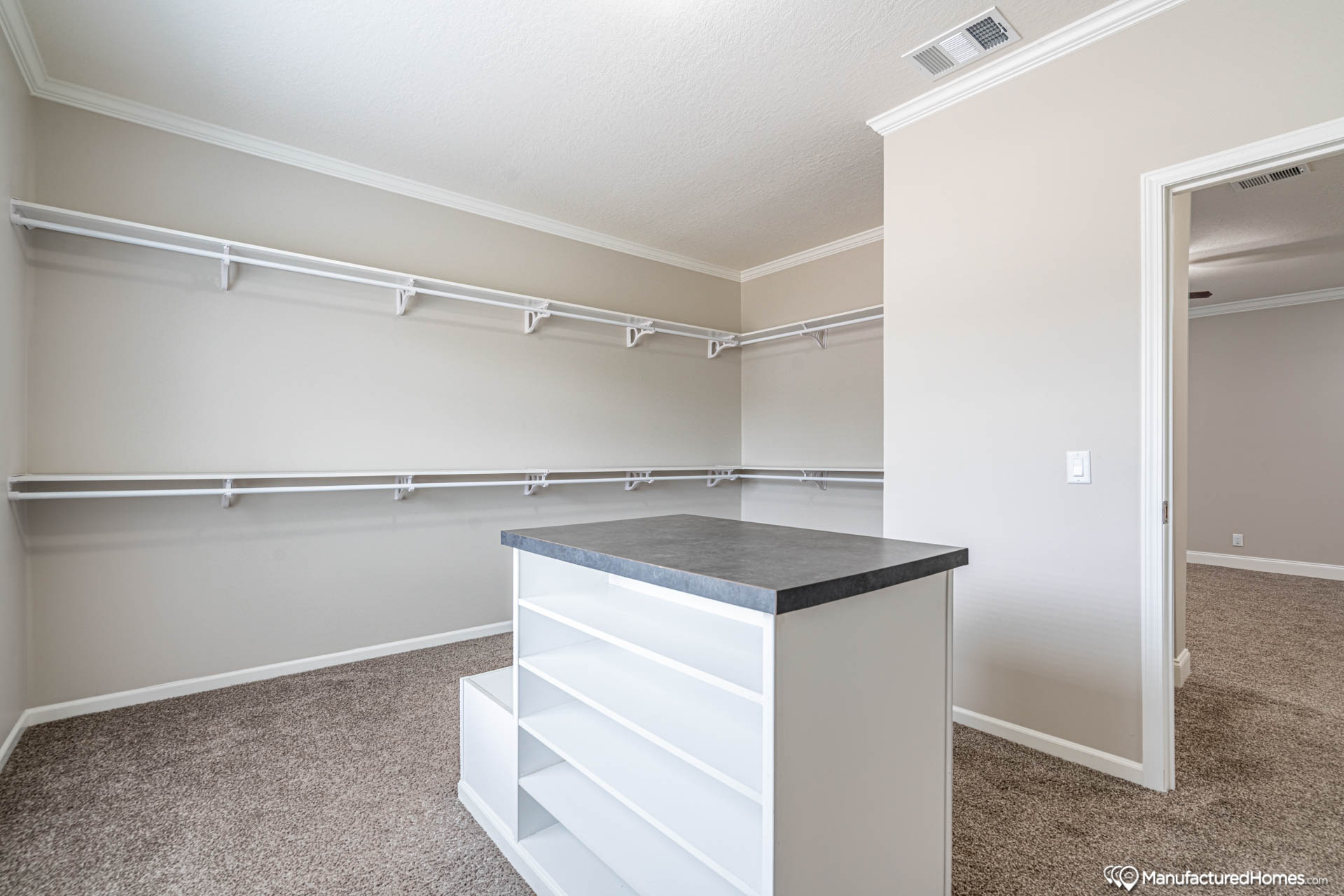 Spacious walk-in closet with beige walls and carpeted floor, featuring multiple white shelves and hanging rods. An island with open shelving is centered, creating a neat and organized feel.