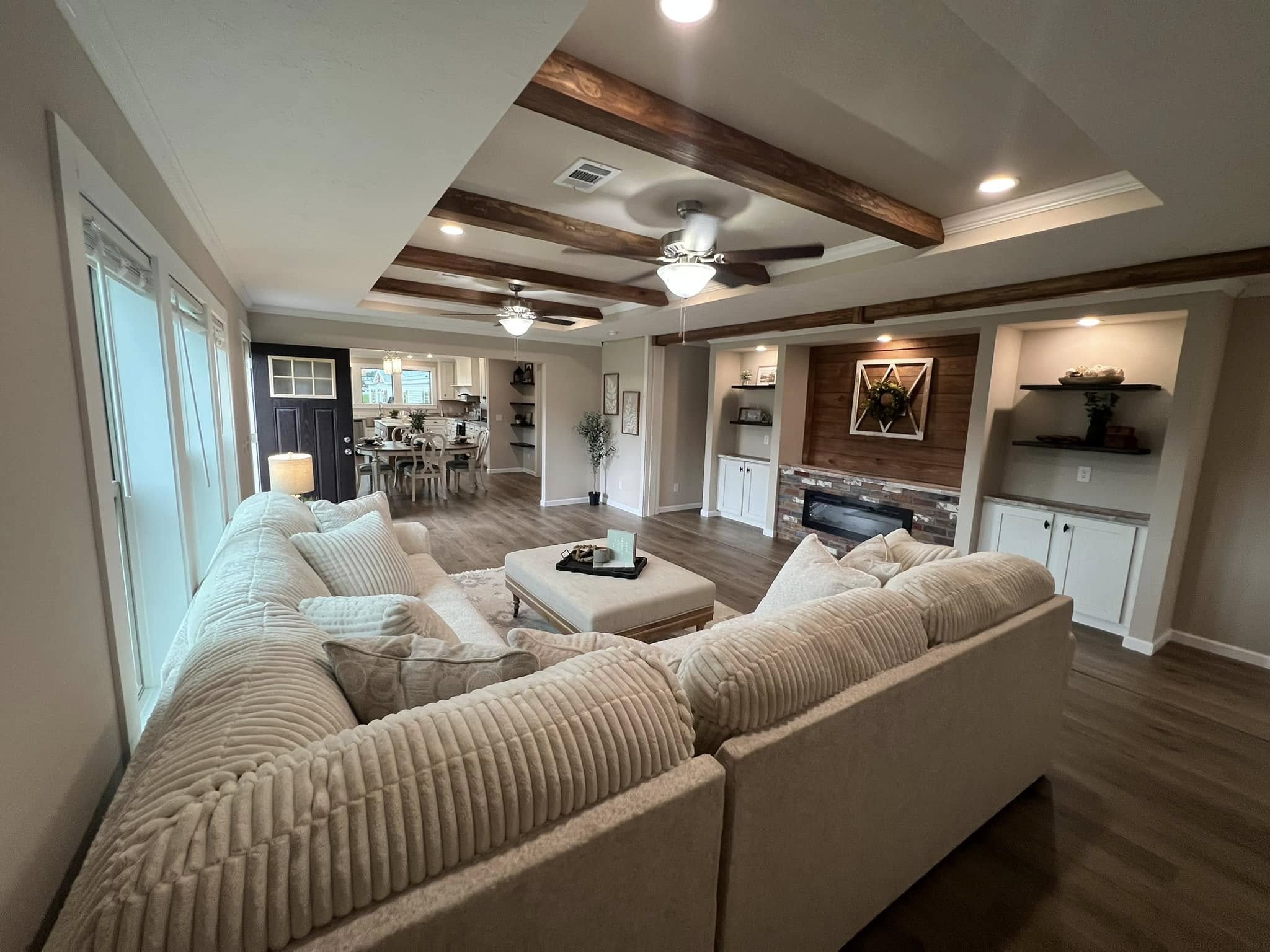 Spacious living room with beige sectional, wooden ceiling beams, and cozy fireplace. Open layout leads to a dining area, creating a warm, inviting atmosphere.