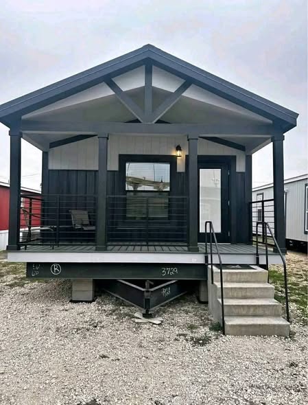 Small modern prefab house with a dark wooden porch, white siding, and black trim. Concrete steps lead to the entrance. Overcast sky sets a calm tone.