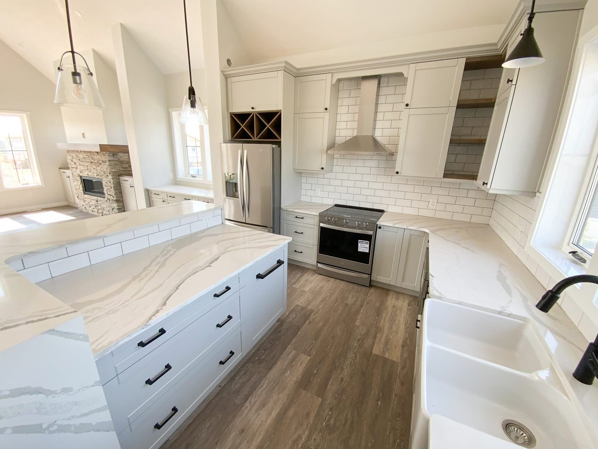 Modern kitchen with white cabinets, marble countertops, and stainless steel appliances. Pendant lights hang above a large island. Warm, inviting tone.