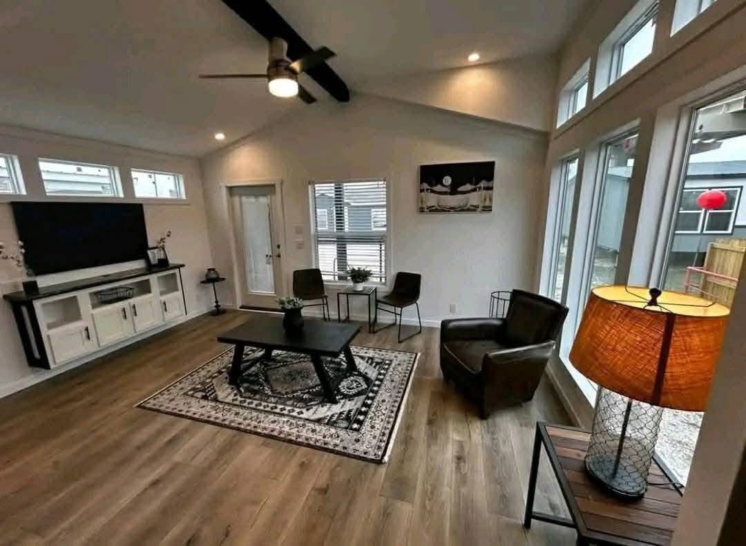 Modern living room with wooden floor, beige walls, large windows, a ceiling fan, wall-mounted TV, and cozy furniture on a patterned rug; warm ambiance.