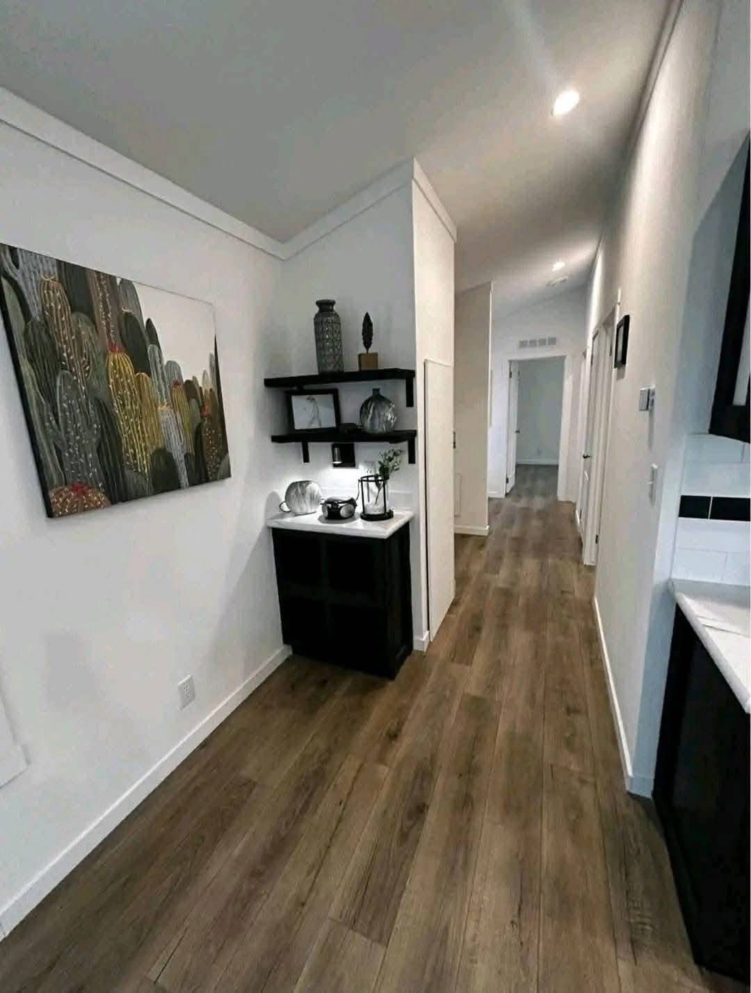 A modern hallway with wooden flooring, white walls, and recessed lighting. Decor includes a cactus painting and shelves with vases and a teapot.