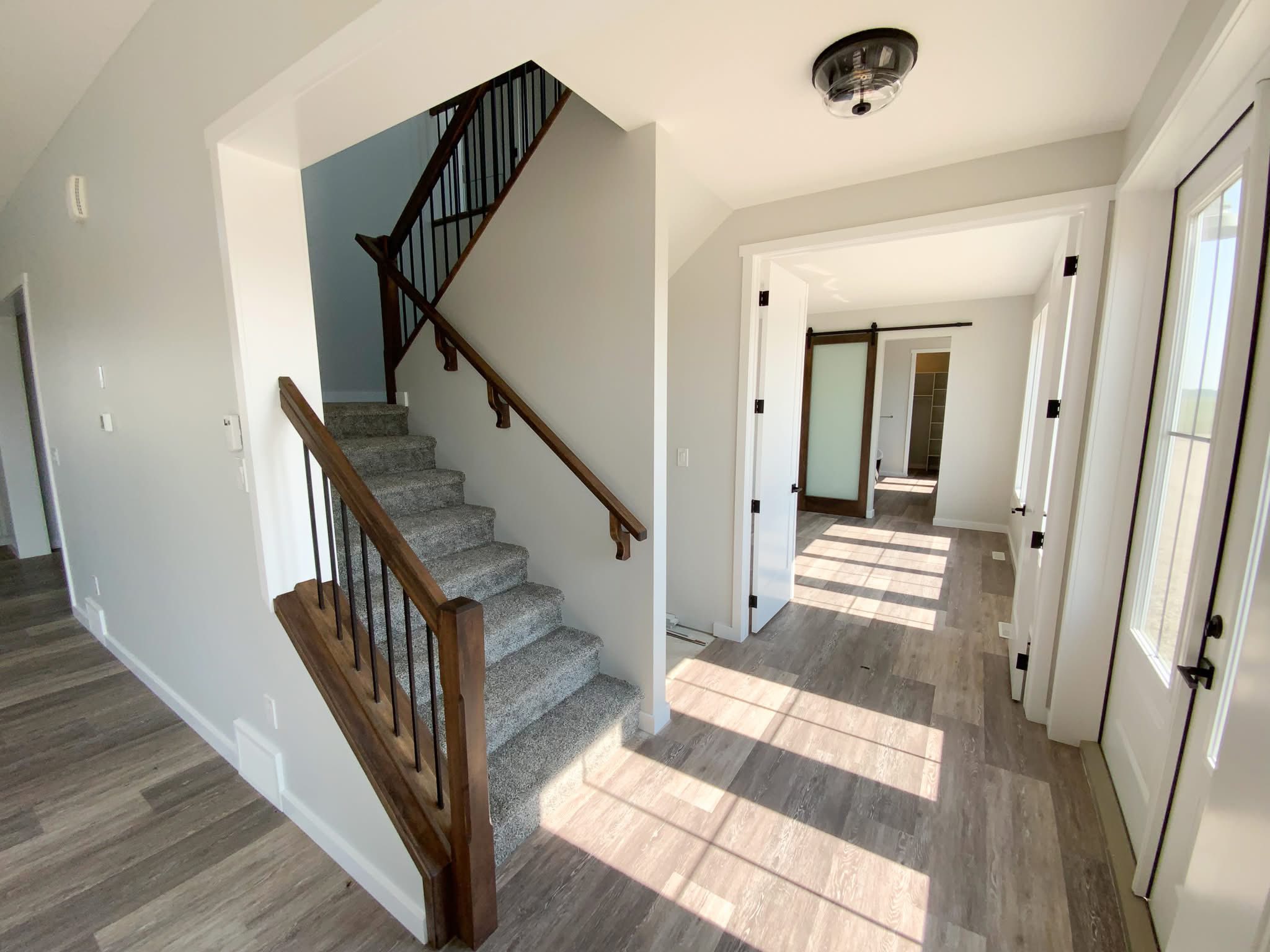 Bright, modern hallway with wooden flooring, carpeted stairs with dark wooden railings, a sliding barn door, and sunlight streaming in through glass doors.
