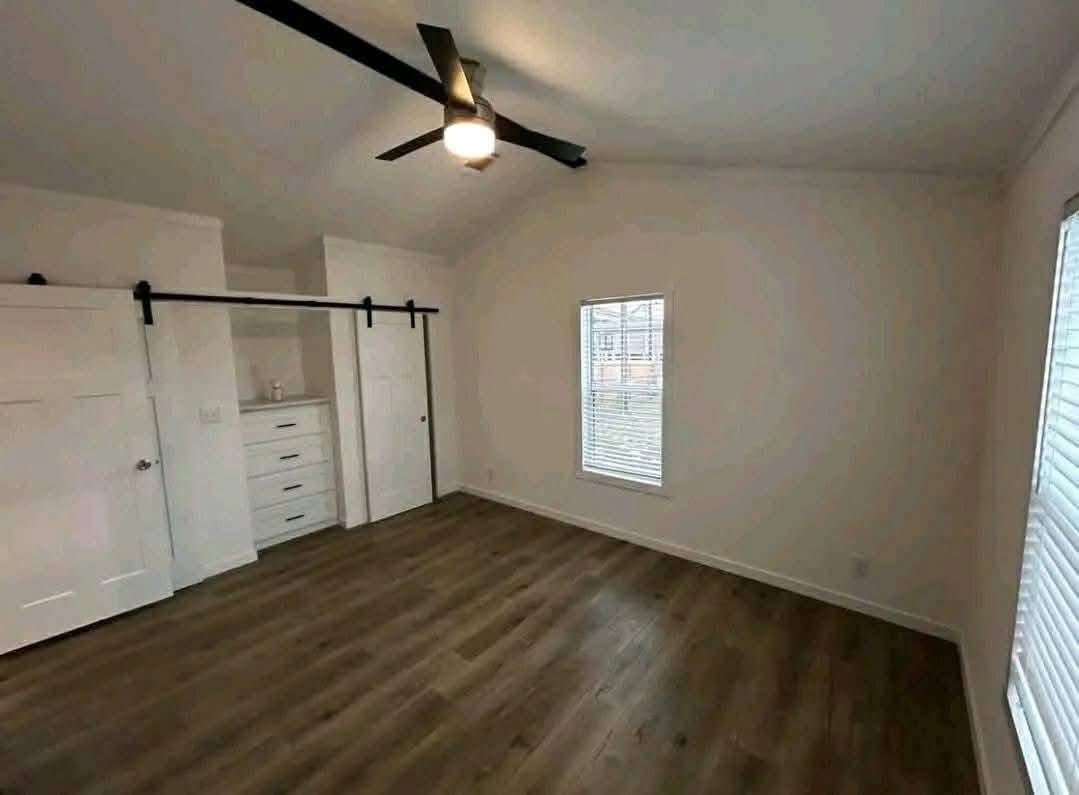 Spacious empty room with white walls, wood flooring, ceiling fan, sliding closet doors, dresser, and large windows letting in natural light.