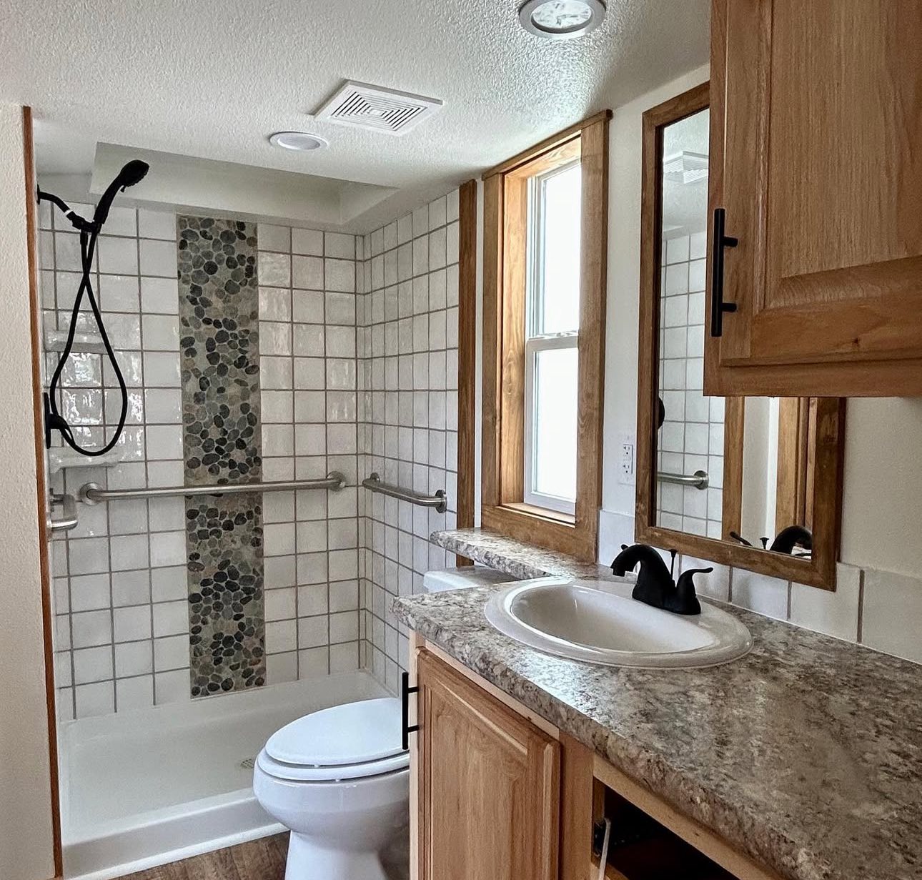A compact, modern bathroom featuring a white tiled shower with a pebble accent strip and stainless steel grab bar. Wooden cabinets with a granite countertop and a black faucet are next to a window, creating a cozy, inviting atmosphere.
