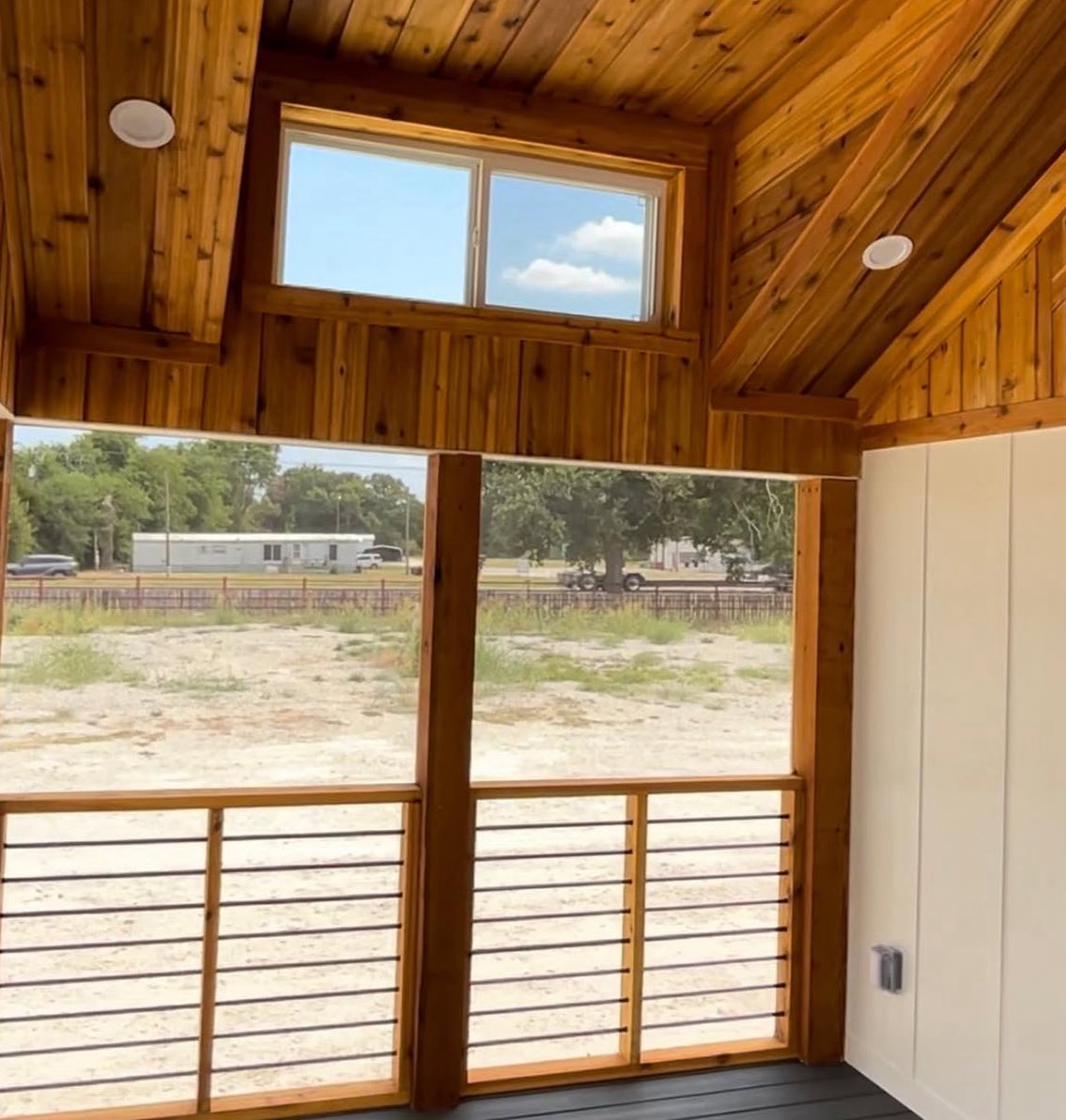 Wood-paneled cabin interior with large windows overlooking a sandy yard and distant trees. The scene feels warm and inviting, with natural light.