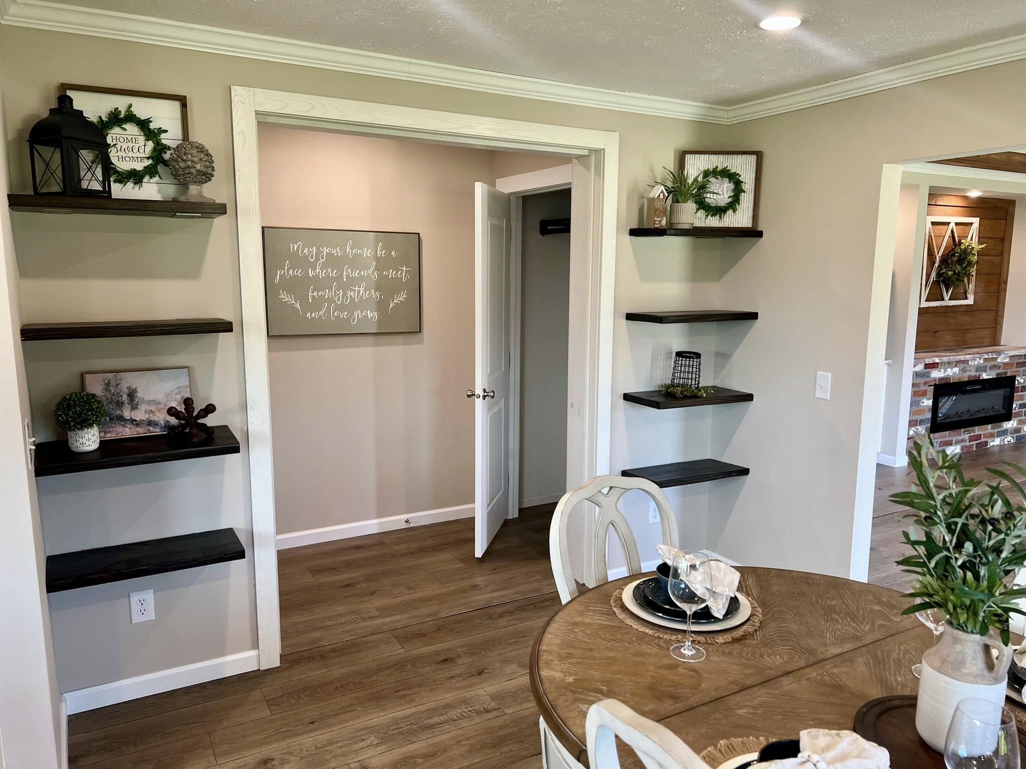 A cozy living space with beige walls features dark wooden floating shelves adorned with greenery and decor. A round wooden table is set for a meal, creating a welcoming atmosphere.