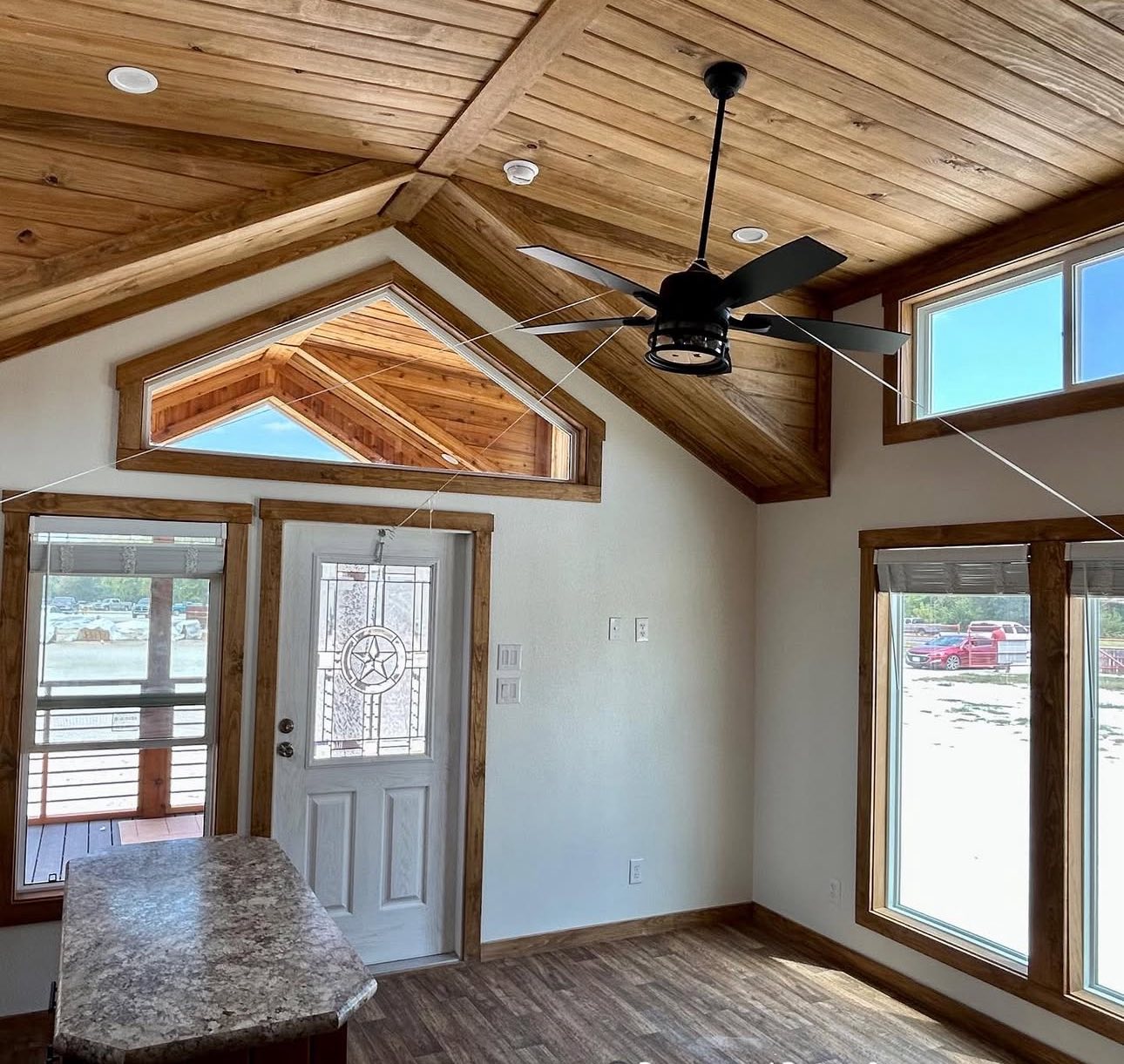 Cozy room with wooden ceiling and natural light. Features a ceiling fan, large windows, and a decorative white door, creating a warm, inviting atmosphere.