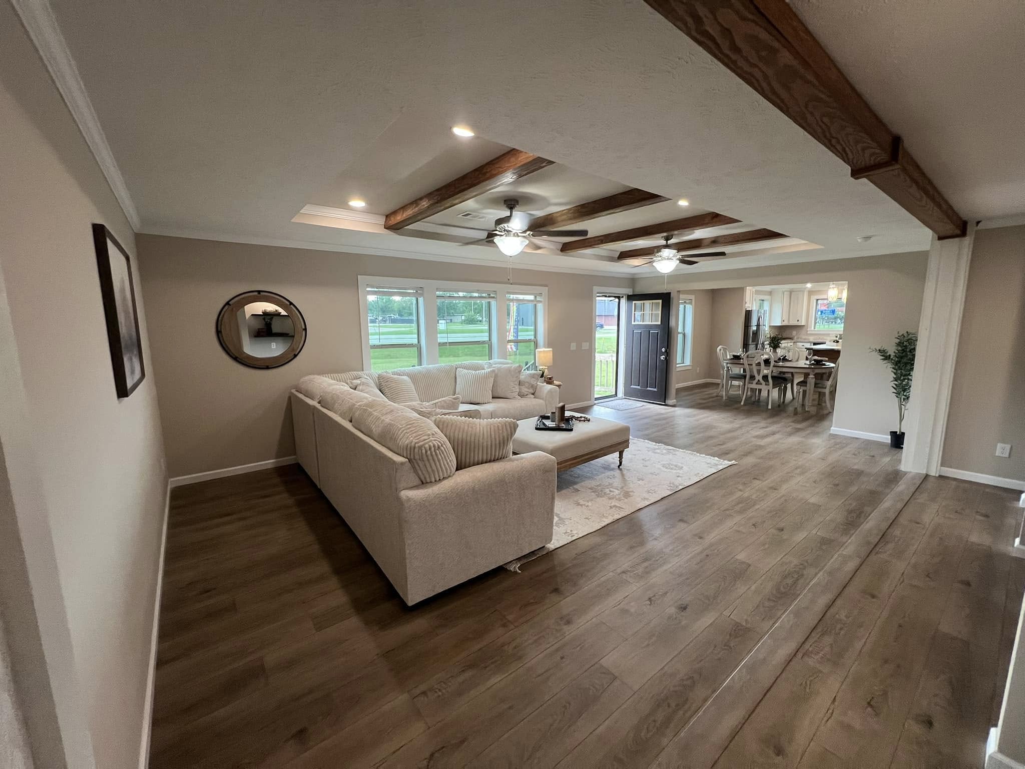 Spacious, modern living room with wood beams, hardwood floors, large sectional sofa, and wall mirror. Bright, inviting with natural light from windows.