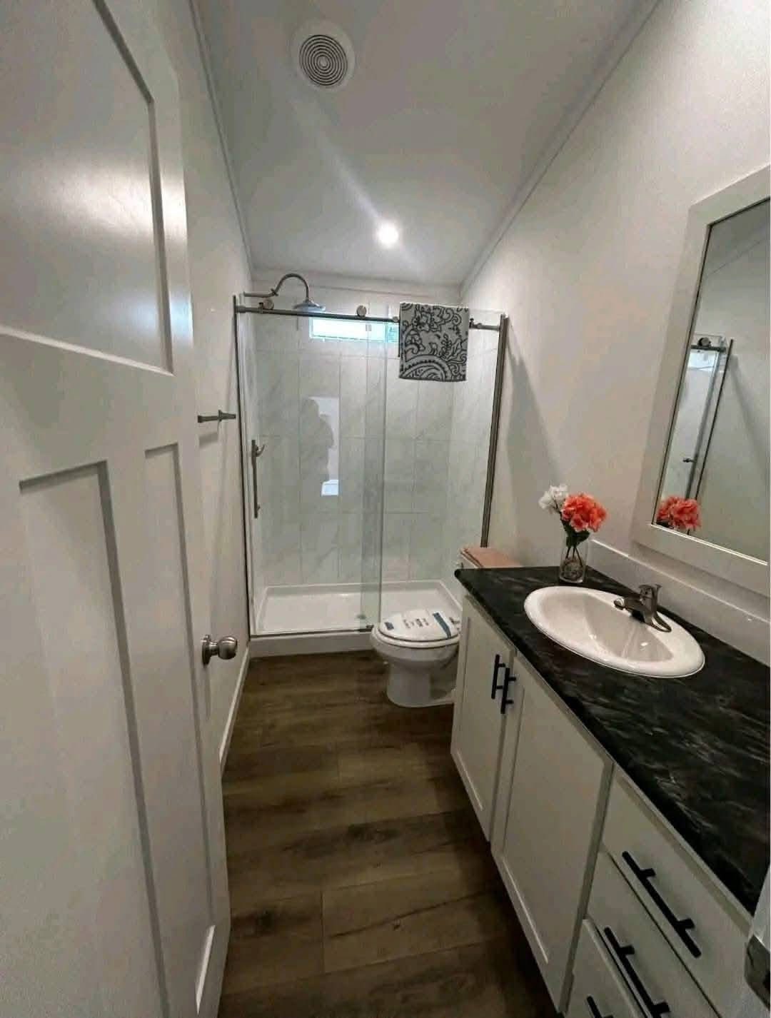 A modern bathroom with wood flooring, a shower with glass doors, white cabinets, a dark countertop, a sink, and a vase with pink flowers.