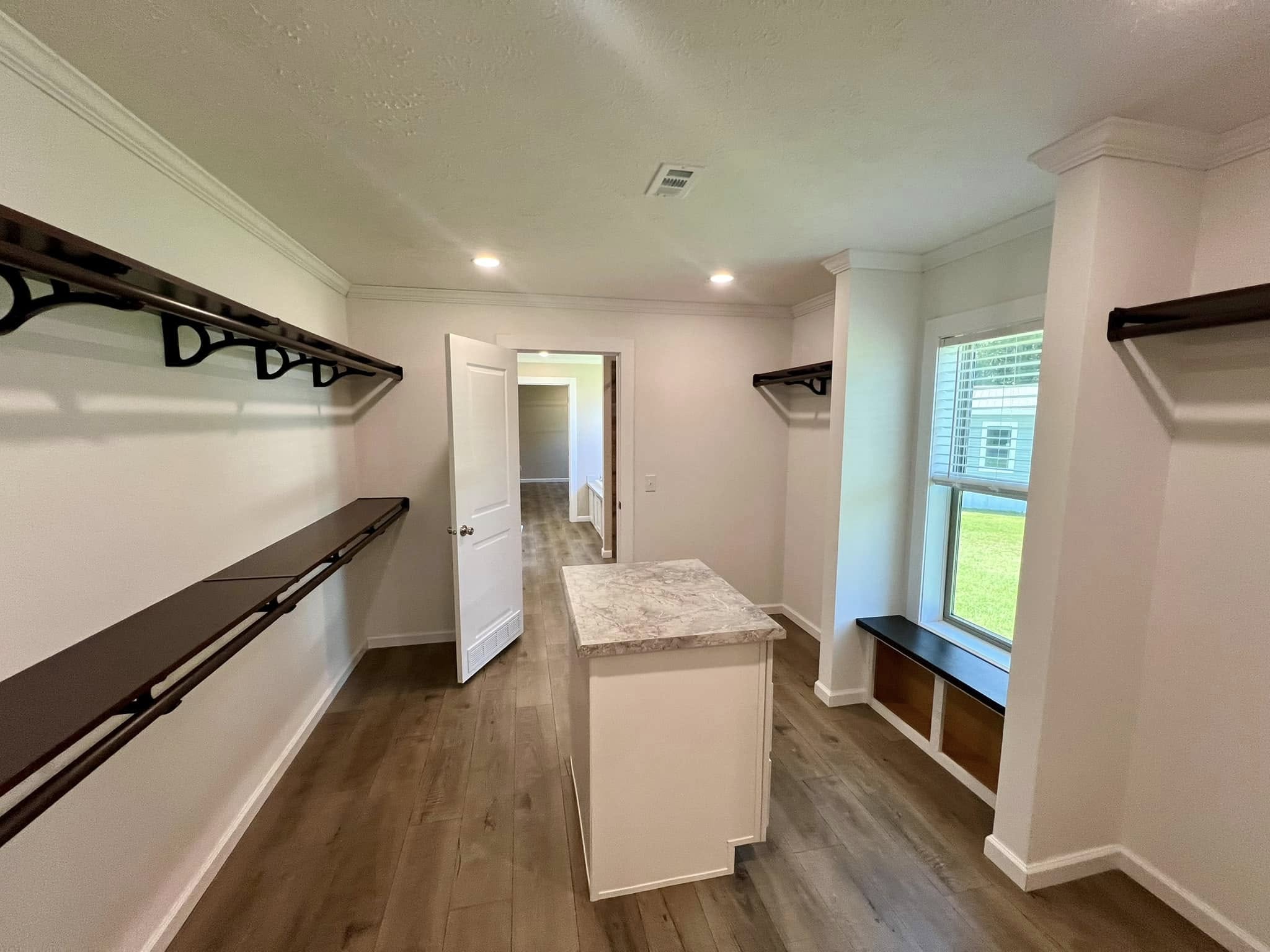 Spacious walk-in closet with wooden shelves and a central island. A window bench with storage offers natural light, creating a modern, airy feel.