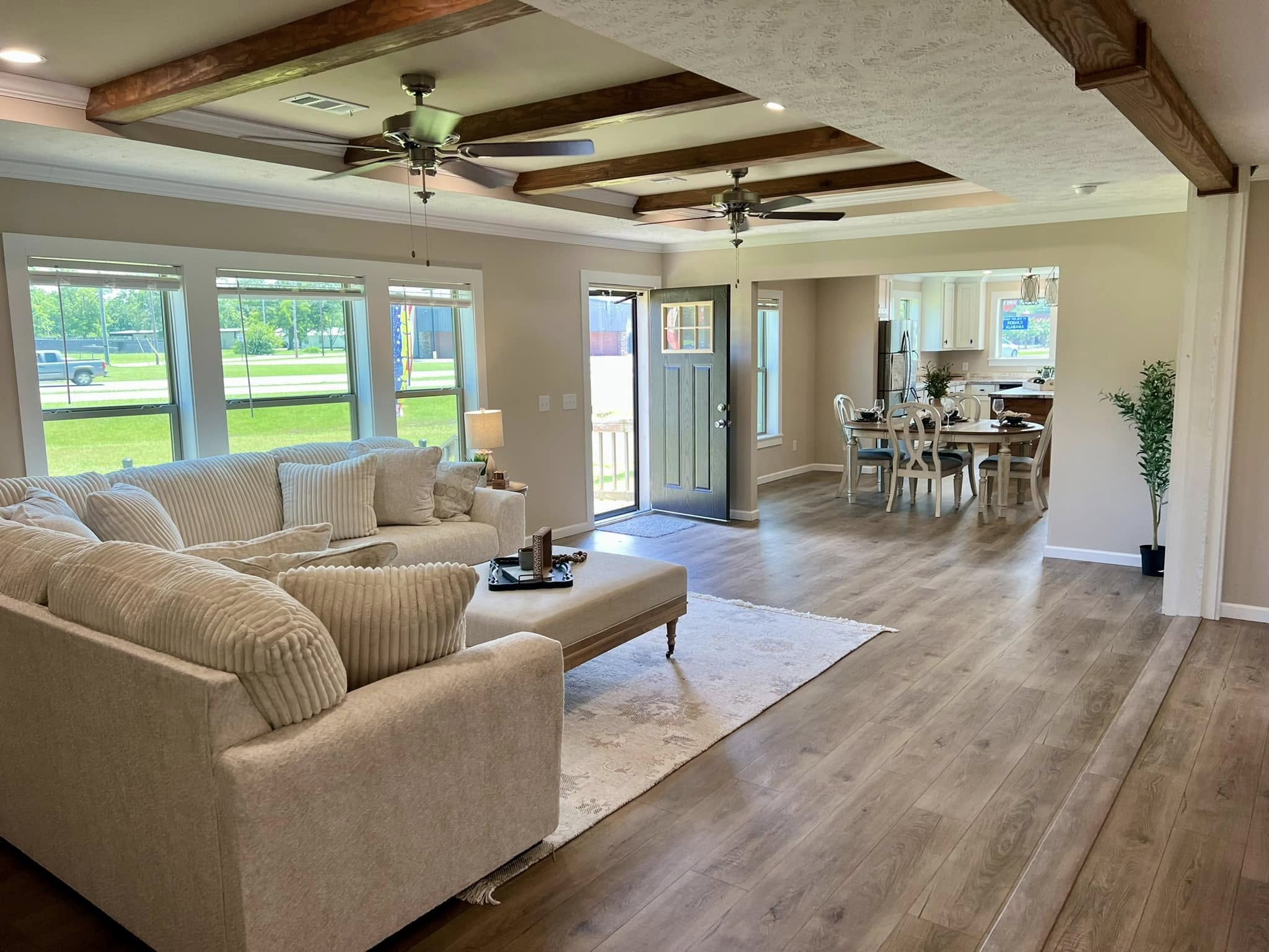 Bright open-plan living room with light wood floors, coffered ceiling, and large windows. Beige sectional sofa and dining area create a cozy, welcoming atmosphere.