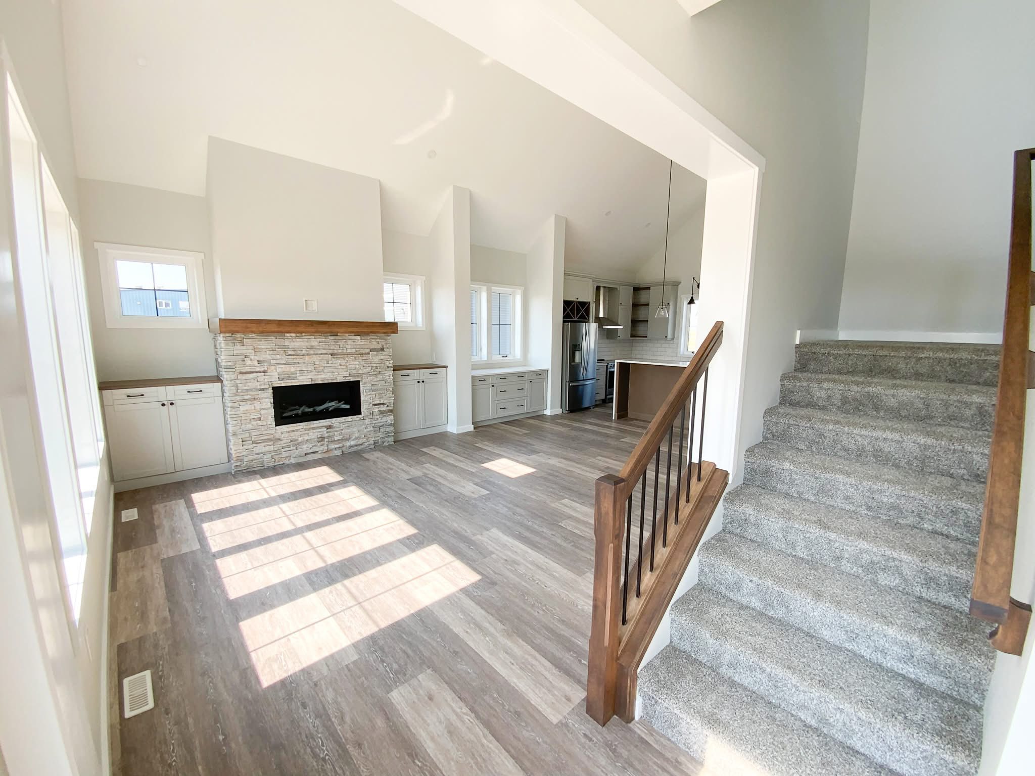 Bright, modern living room with stone fireplace, large windows, light wood flooring, and a gray carpeted staircase leading upstairs, evoking a cozy ambiance.