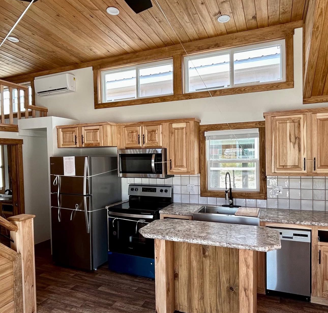 Cozy kitchen with wooden cabinets, stainless steel appliances, and marble countertops. Large windows emit natural light, creating a warm atmosphere.