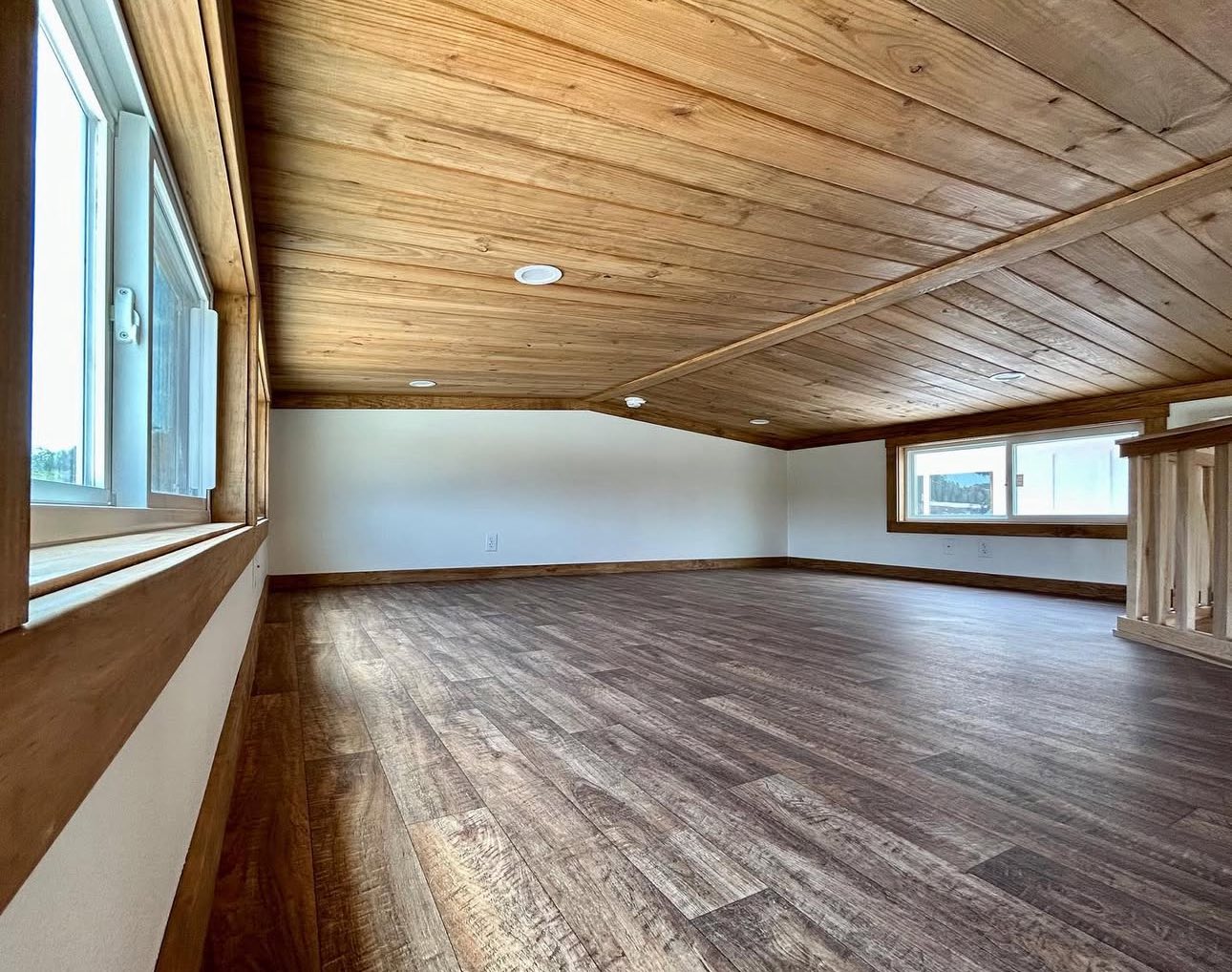 A cozy, rustic attic with wooden floors and ceiling. Two windows allow natural light, creating a warm ambiance. The space feels open and inviting.