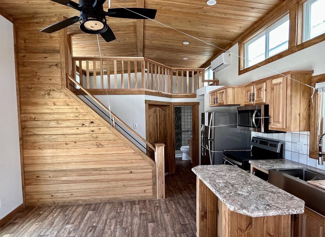 Warmly lit kitchen with wooden cabinets, lofted ceiling, and staircase. Modern appliances, granite counters, and ceiling fan create a cozy, rustic vibe.