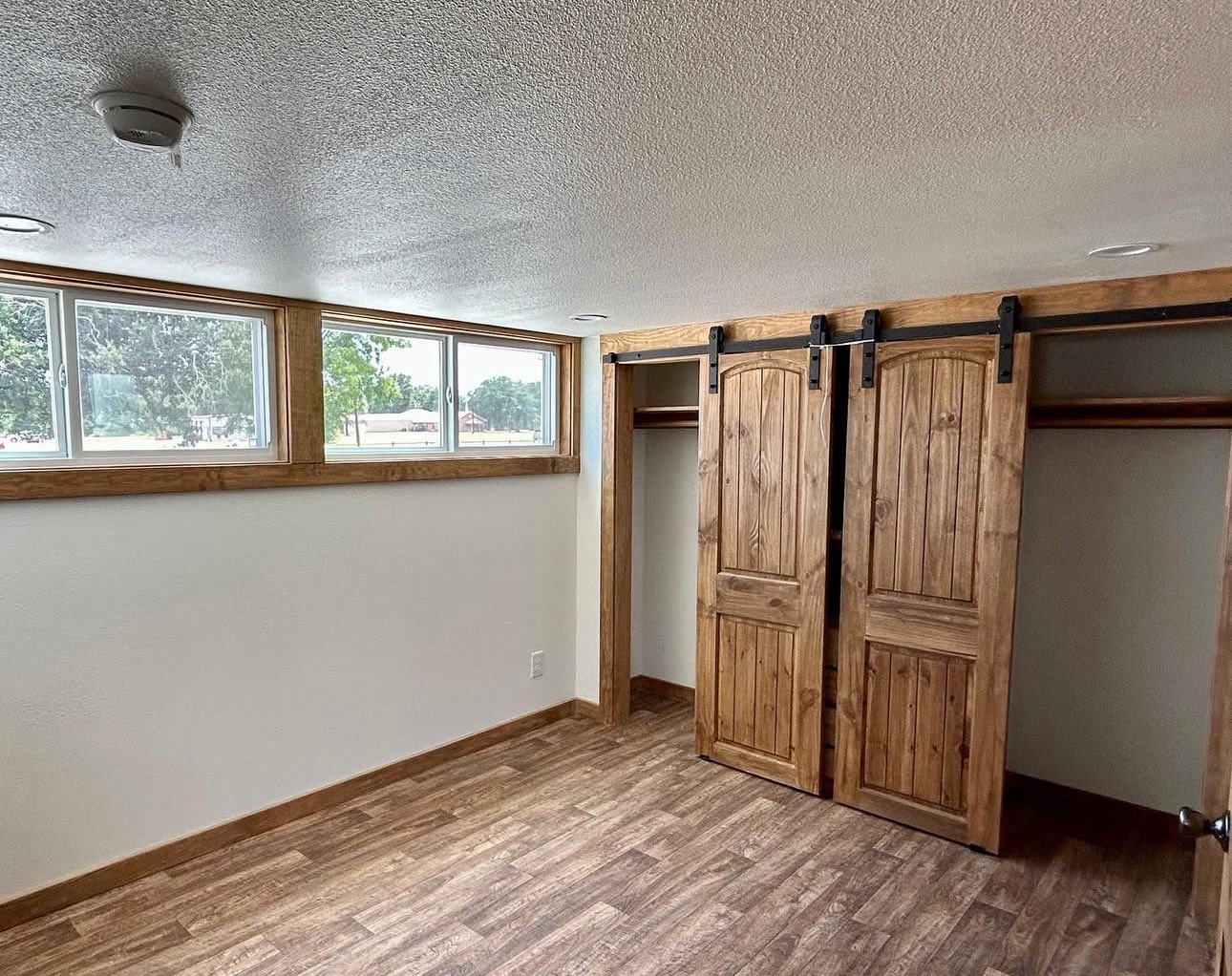 A sparsely furnished room with a wooden floor, a row of small windows on the left, and wooden sliding barn doors on the right hiding a closet space. The atmosphere is simple and rustic.