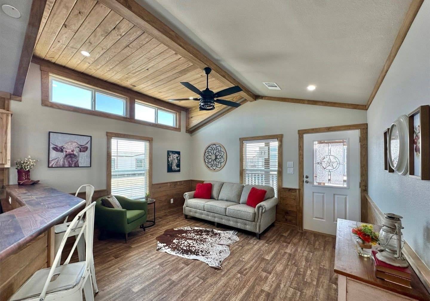 Cozy living room with vaulted wooden ceiling, ceiling fan, and natural light. Features a gray sofa, cowhide rug, and rustic decor on wooden floors.