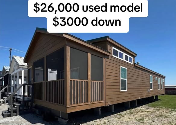 Brown mobile home with screened porch, large windows, and gable roof under clear blue sky. Text overlay: "$26,000 used model, $3,000 down."