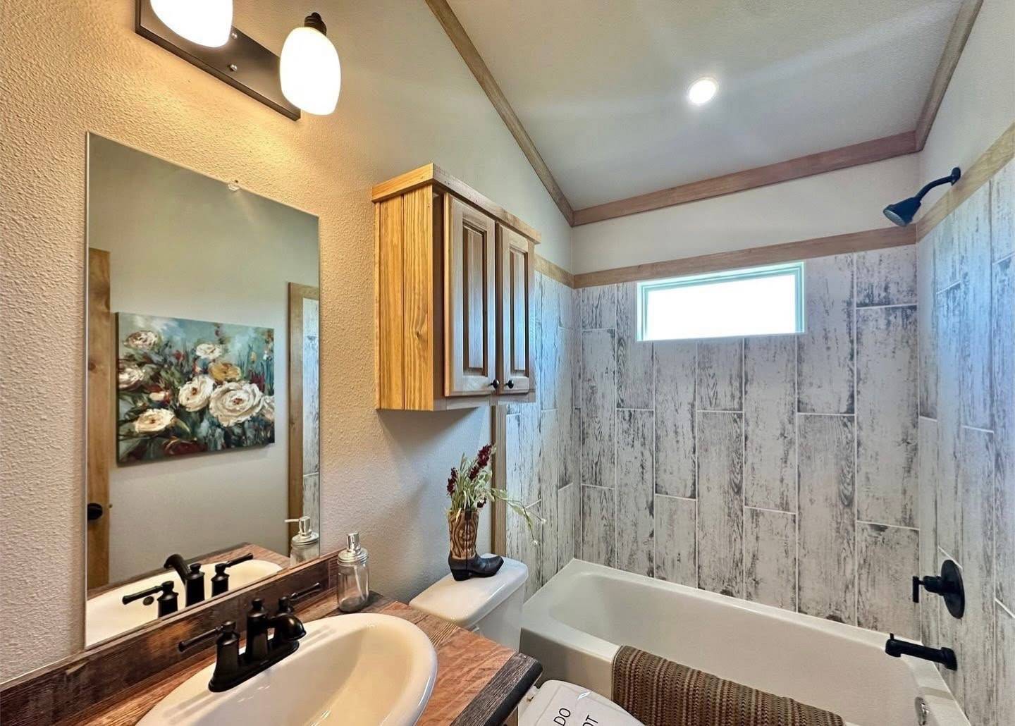 Bathroom with rustic decor: wooden vanity, floral painting, and wood-paneled bathtub area. Soft lighting and warm tones create a cozy ambiance.