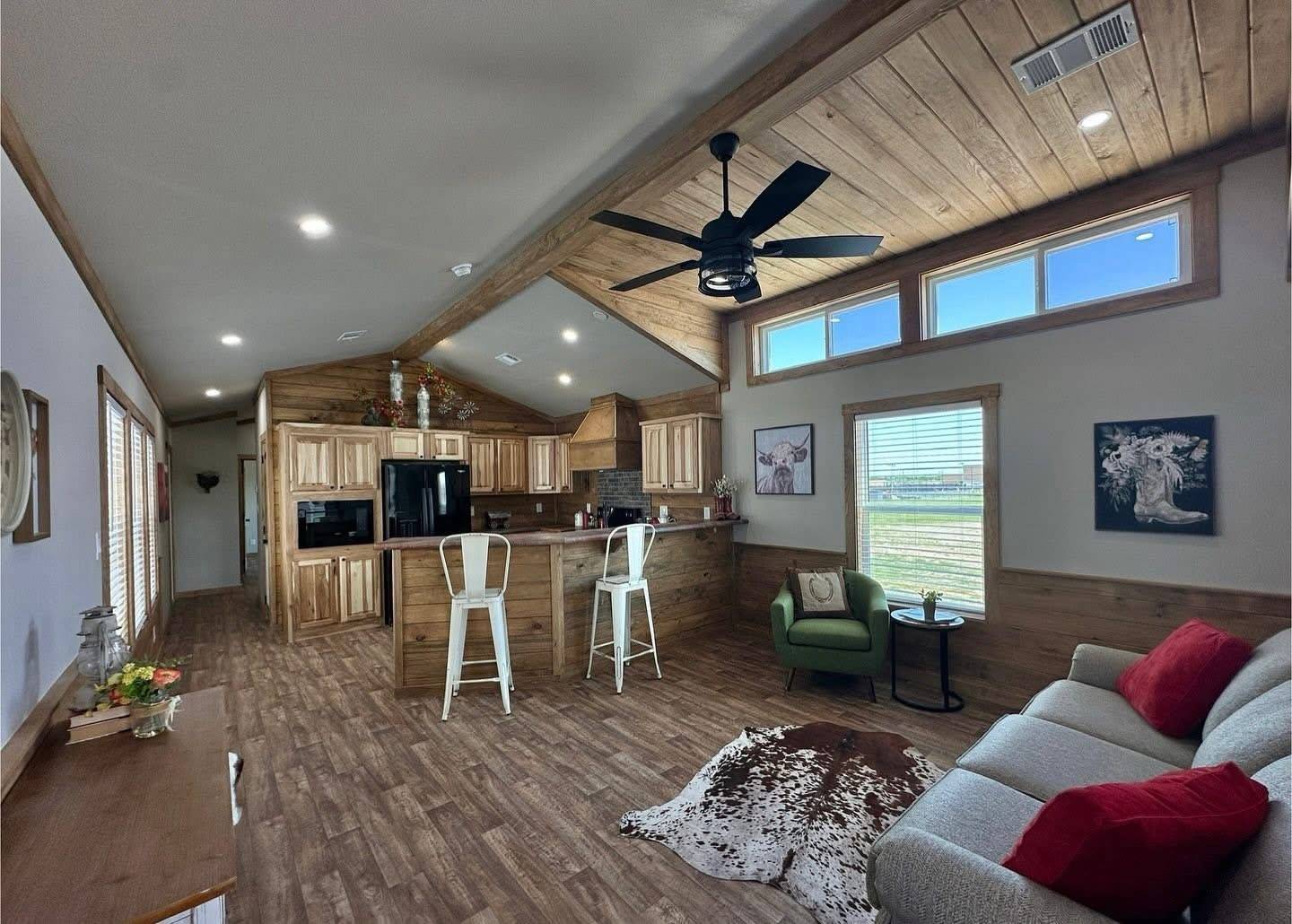 Cozy living area with wooden ceiling and floors. Features include a rustic kitchen, a ceiling fan, sofa with red pillows, and a cowhide rug. Natural light streams in.