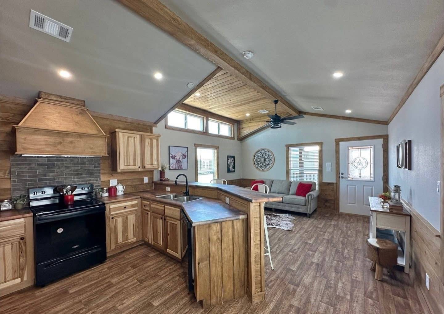 Cozy open-concept living room and kitchen with wood accents and flooring. Features a black stove, sectional sofa, and decorative wall art. Bright and inviting.
