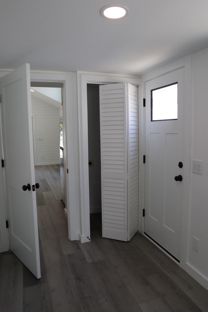 A small, white hallway with three doors: one open, a louvered closet door, and an entrance door with a window. Dark handles contrast bright walls. Modern and minimal.