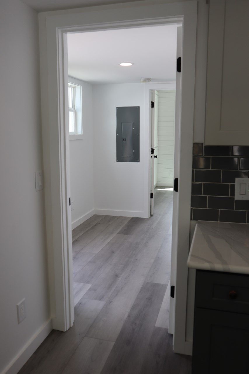 View from a kitchen with gray tiled backsplash and marble countertop into a bright hallway with light wood flooring and a utility panel.