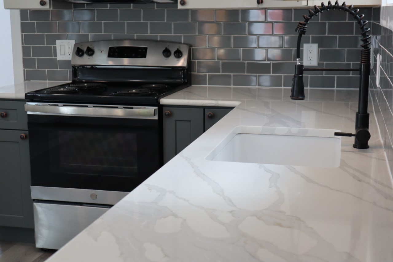 Modern kitchen with sleek white marble countertops, dark cabinets, and gray subway tile backsplash. Features include a black faucet and stainless steel stove.