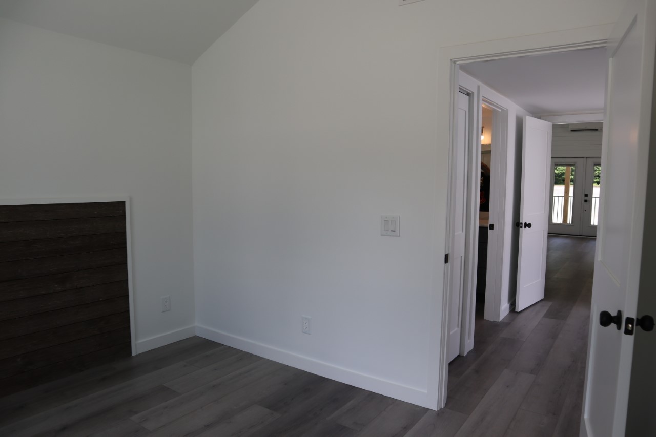 A bright, empty room with white walls and dark wood flooring. Two open doors lead to a hallway with natural light, creating a clean, modern feel.
