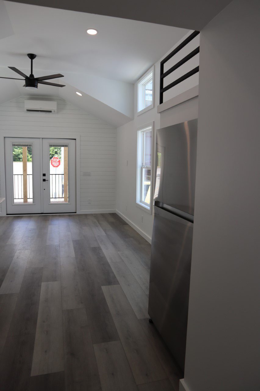 Minimalist, well-lit kitchen with high ceiling, wooden floors, stainless steel fridge, and double glass doors opening to greenery, creating an airy feel.