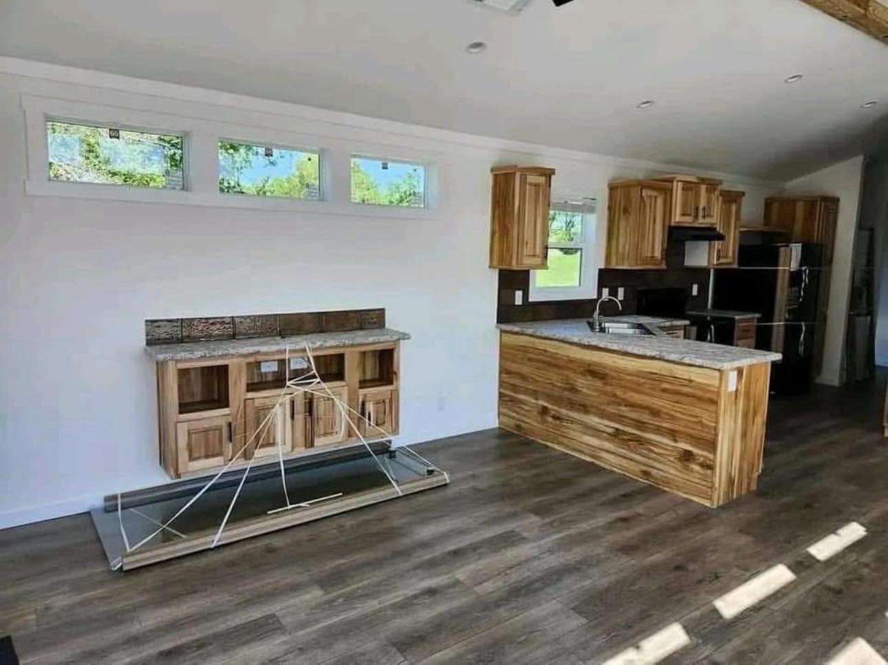Modern kitchen with wooden cabinetry, gray countertops, and large windows. A wooden cabinet with taped string lines is on the left. Sunlight filters in, creating a warm, inviting atmosphere.