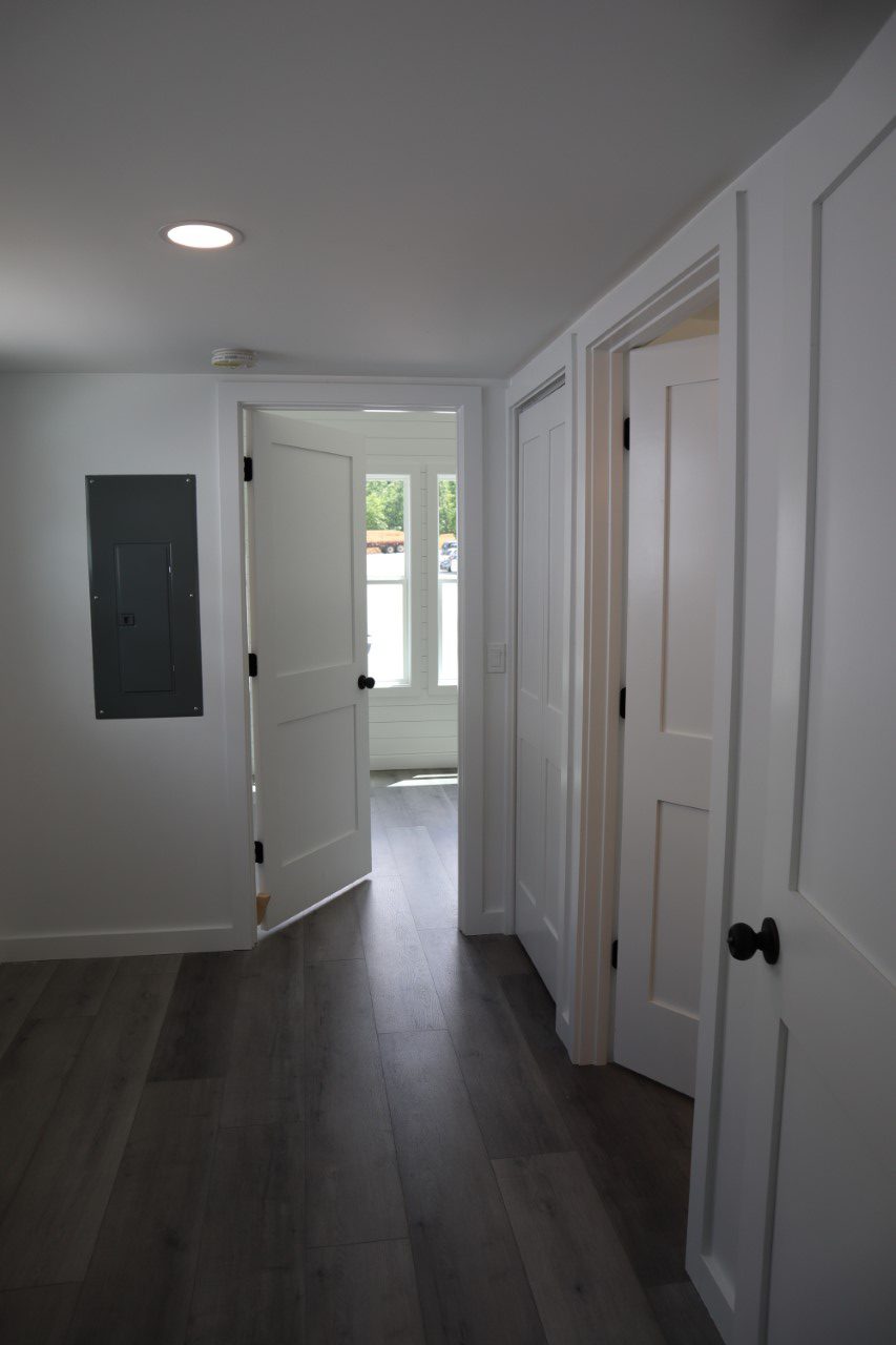 A hallway with dark wood flooring and white walls. Three doors with black knobs are visible, one partially open. Sunlight streams in through a window.