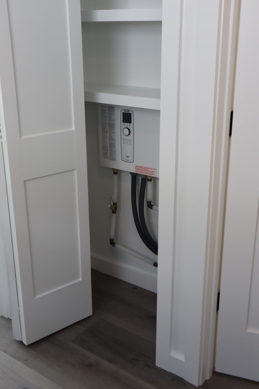 Small white closet with an open door revealing a wall-mounted tankless water heater. The interior is minimal, with pipes visible below the unit.