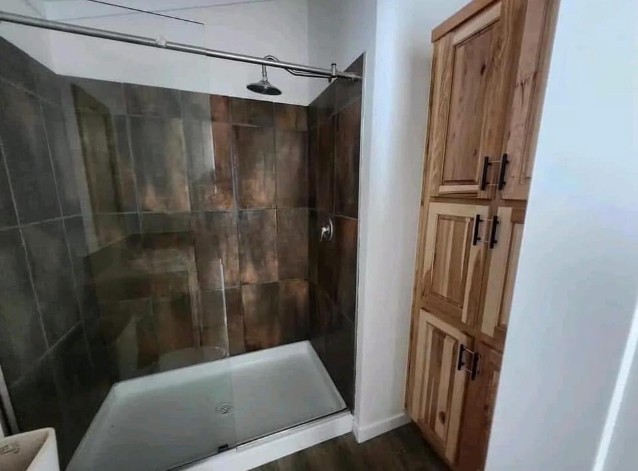 A modern bathroom with a glass-enclosed shower featuring dark brown tiles. To the right, there is a tall, wooden cabinet with black handles.